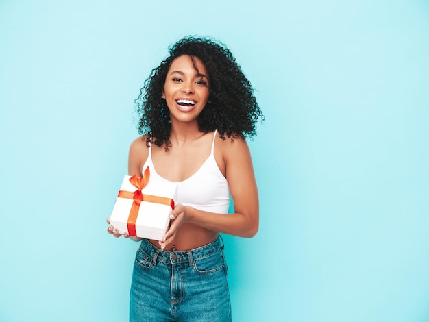 Beautiful black woman with afro curls hairstyle Smiling model dressed in white summer clothes Sexy carefree female posing near blue wall in studio Tanned and cheerful Holding gift box Isolated