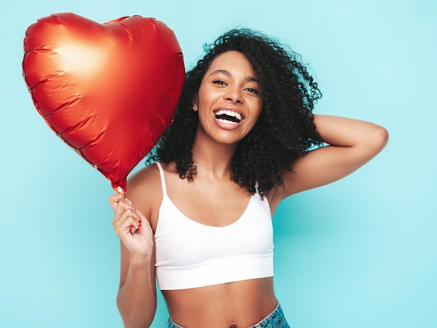 Free photo beautiful black woman with afro curls hairstyle smiling model dressed in white summer clothes sexy carefree female posing near blue wall in studio tanned and cheerful holding heart air balloon