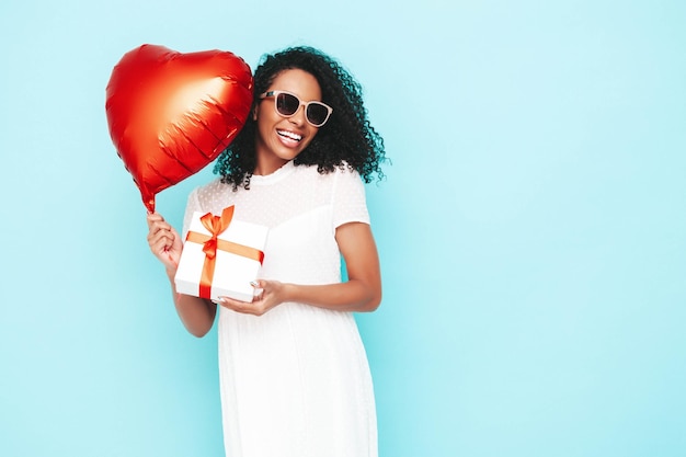 Beautiful black woman with afro curls hairstyle Smiling model dressed in white summer dress Sexy carefree female posing near blue wall in studio Holding heart air balloon and gift box Isolated