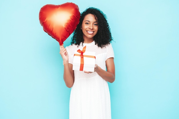 Beautiful black woman with afro curls hairstyle Smiling model dressed in white summer dress Sexy carefree female posing near blue wall in studio Holding heart air balloon and gift box Isolated