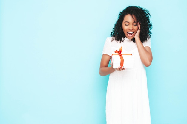 Free photo beautiful black woman with afro curls hairstyle smiling model dressed in white summer dress sexy carefree female posing near blue wall in studio tanned and cheerful holding gift box isolated