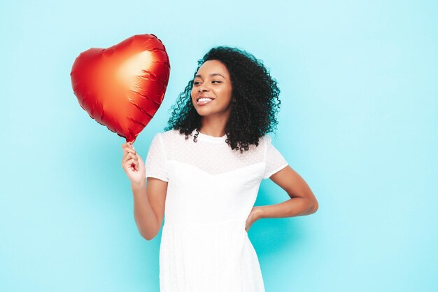 Beautiful black woman with afro curls hairstyle Smiling model dressed in white summer dress Sexy carefree female posing near blue wall in studio Tanned and cheerful Holding heart air balloon