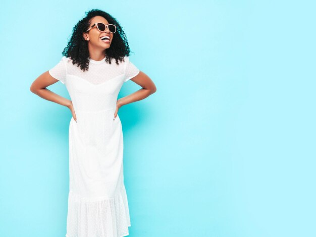 Красивая негритянка с прической афро-кудри Улыбающаяся модель, одетая в белое летнее платье Сексуальная беззаботная женщина позирует возле синей стены в студии Загорелая и веселая в солнцезащитных очках Изолированный
