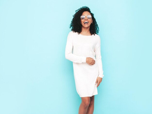 Beautiful black woman with afro curls hairstyle Smiling model dressed in white summer dress Sexy carefree female posing near blue wall in studio Tanned and cheerful in sunglasses Isolated