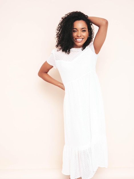 Beautiful black woman with afro curls hairstyle Smiling model dressed in white summer dress Sexy carefree female posing near wall in studio Tanned and cheerful Isolated