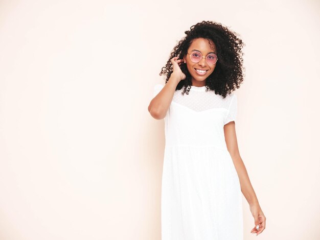 Beautiful black woman with afro curls hairstyle Smiling model dressed in white summer dress Sexy carefree female posing near wall in studio Tanned and cheerful Isolated