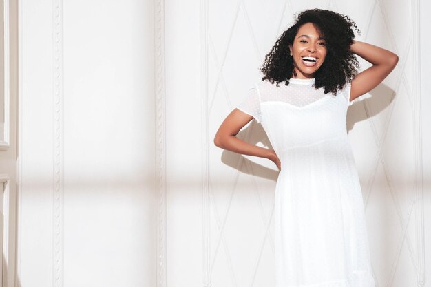 Beautiful black woman with afro curls hairstyle Smiling model dressed in white summer dress Sexy carefree female posing near wall in studio Tanned and cheerful At sunny day Shadow from window
