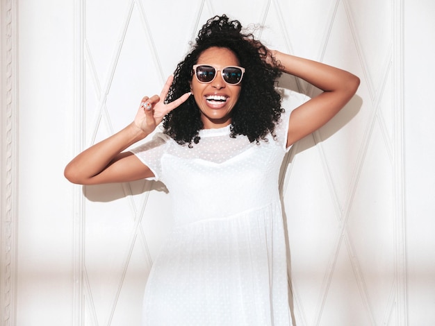 Beautiful black woman with afro curls hairstyle Smiling model dressed in white summer dress Sexy carefree female posing near wall in studio Tanned and cheerful At sunny day Shadow from window