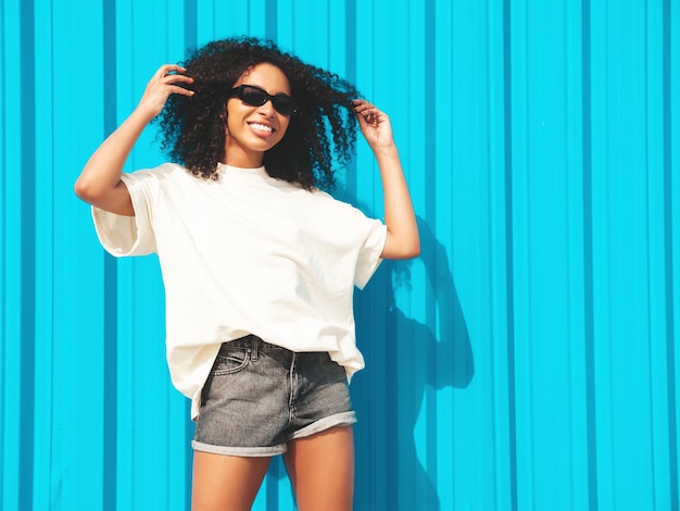 Красивая негритянка с прической афро-кудриУлыбающаяся хипстерская модель в белой футболке Сексуальная беззаботная женщина позирует на улице возле синей стены в солнцезащитных очках Веселая и счастливая