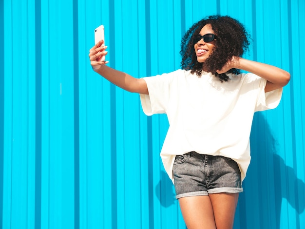 Beautiful black woman with afro curls hairstyleSmiling hipster model in white tshirt Sexy carefree female posing in the street near blue wall in sunglasses Cheerful and happyTaking selfie photo