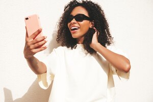 Beautiful black woman with afro curls hairstylesmiling hipster model in white tshirt sexy carefree female posing in the street near white wall in sunglasses cheerful and happytaking selfie photo