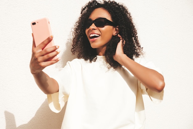 Beautiful black woman with afro curls hairstyleSmiling hipster model in white tshirt Sexy carefree female posing in the street near white wall in sunglasses Cheerful and happyTaking selfie photo