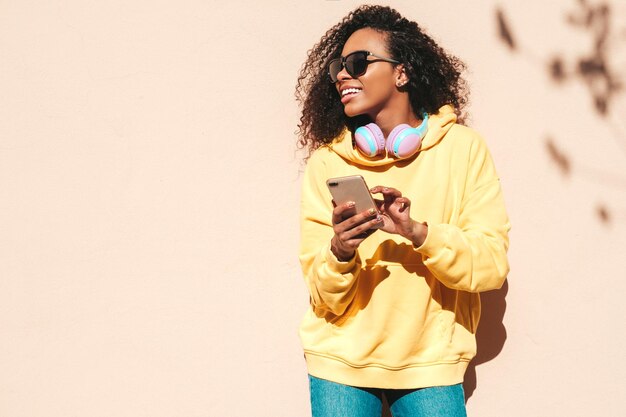 Beautiful black woman with afro curls hairstyleSmiling model in yellow hoodie Sexy carefree female posing on the street background in sunglasses Looking at smartphone screen using apps