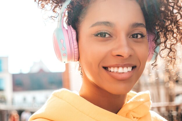 Beautiful black woman with afro curls hairstyleSmiling model in yellow hoodieSexy carefree female enjoying listening music in wireless headphonesPosing on street background at sunset