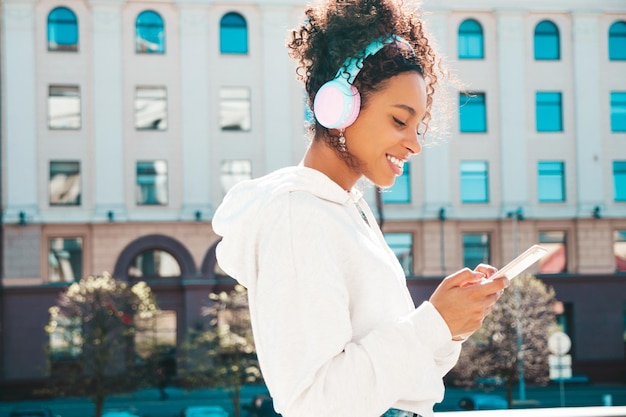 Beautiful black woman with afro curls hairstyleSmiling model in yellow hoodieSexy carefree female enjoying listening music in wireless headphonesPosing on street background at sunsetHolds phone