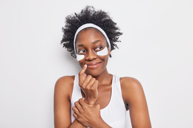 아름다운 흑인 젊은 여성은 꿈결 같은 만족스러운 표정을 보이며 입술 모서리 근처에 손가락을 유지하고 다크 서클 치료를 위한 패치를 적용합니다