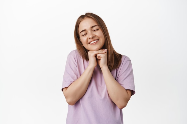 Beautiful blond girl close eyes, smiling and dreaming, praying or making wish, anticipating smth beautiful, daydreaming, standing against white background in t-shirt
