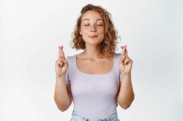 Beautiful blond girl praying, making wish with crossed fingers and closed eyes, wants to achieve something on white.