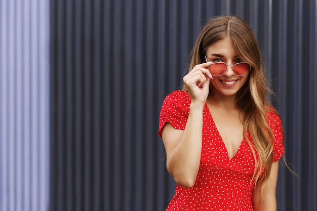 赤いサングラスと夏のドレスの美しいブロンドの女の子がカメラに微笑んで、色付きの壁の近くの晴れた日に屋外に立っています。