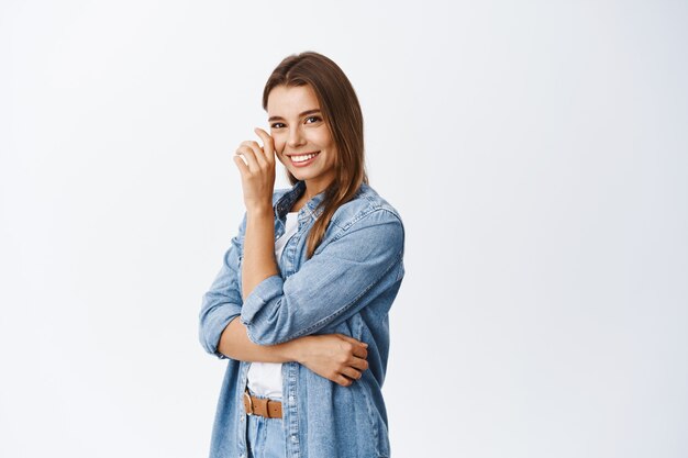 Beautiful blond girl touching her face and smiling white teeth, standing half turned against white wall in casual clothes