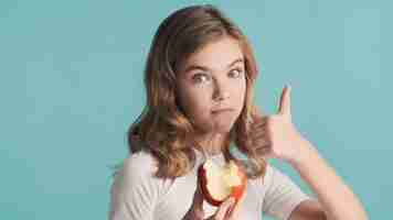 Free photo beautiful blond teenager girl with wavy hair eating delicious apple keeping thumb up over blue background. like it