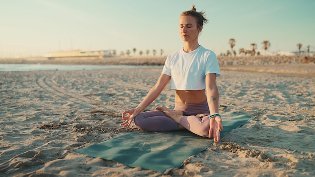 Beautiful blond woman dressed in sportswear sitting in lotus pose by the sea Yogi girl meditating on mat on beach