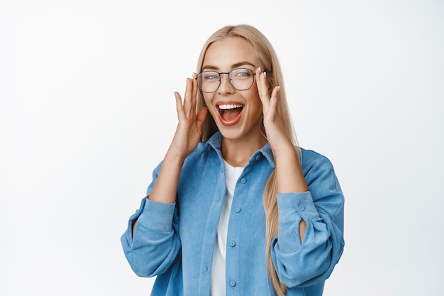Beautiful blond woman put on glasses, laughing and smiling with happy face expression, standing in blue shirt on white