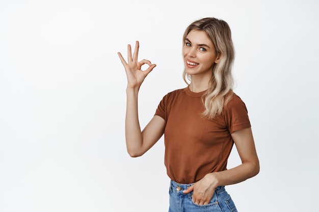 Beautiful blond woman shows okay, OK sign and smiling, giving approval, recommending something good, standing on white