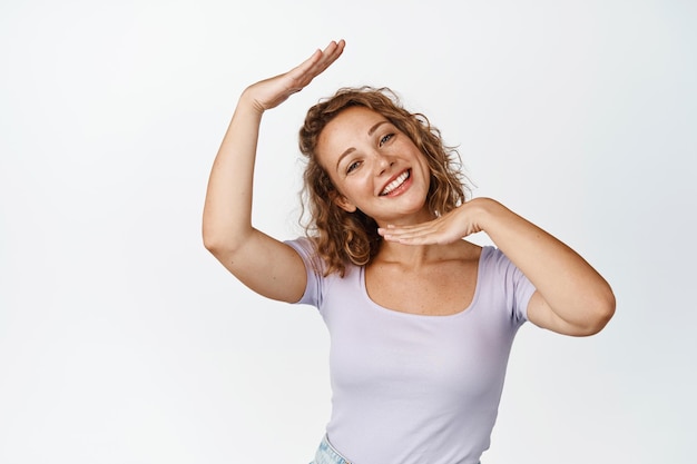 Beautiful blond woman with curly hair, shows box gesture on her face, smiling and looking cute at camera, standing in tshirt over white background.