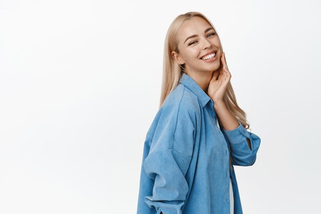 Beautiful blond woman with glowing healthy skin smiling happy at camera touching her face enjoy softness standing over white background