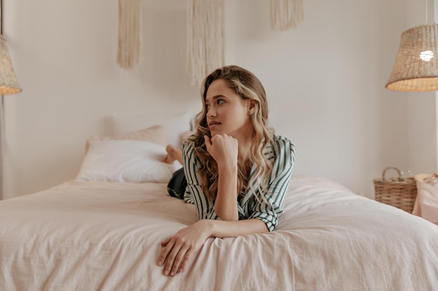 Beautiful blonde curly woman in striped stylish shirt rests on soft big bed Charming lady in pajam