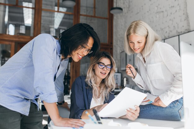 Beautiful blonde girl in glasses showing report to asian colleague which standing beside her table