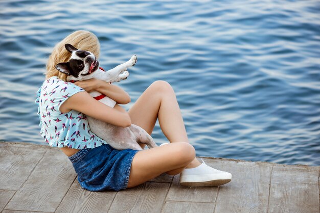 Beautiful blonde girl sitting, playing with French bulldog near sea