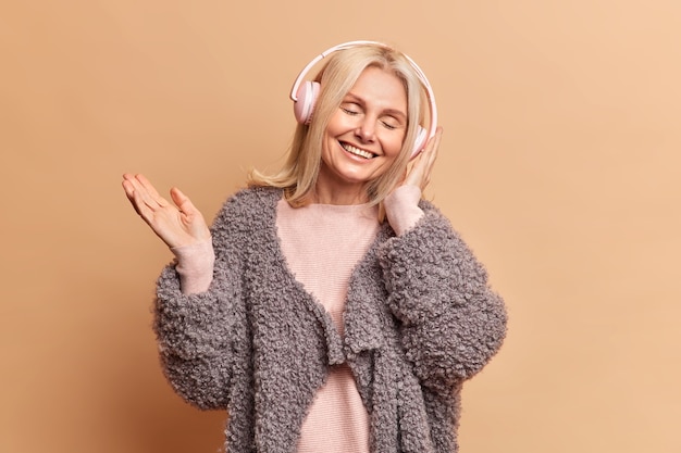 Beautiful blonde middle aged woman closes eyes and wears stereo headphones listens pleasant melody via headphones dressed in fashionable winter clothes isolated over brown studio wall