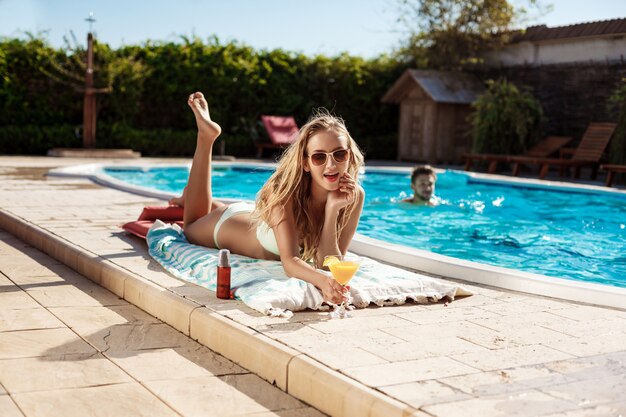 Beautiful blonde woman drinking cocktail, sunbathing, lying near swimming pool
