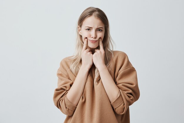 beautiful blonde woman making grimace, touching her cheeks with fingers, making herself smile, frowning face, being displeased and upset. Face expression and negative emotions