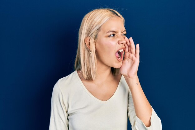 Beautiful blonde woman wearing casual sweater shouting and screaming loud to side with hand on mouth communication concept