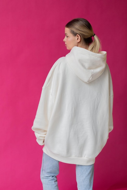Beautiful blonde woman in a white hoodie and blue jeans posing on a pink background