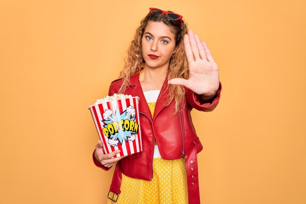 Beautiful blonde woman with blue eyes eating salty popcorn snack over yellow background with open hand doing stop sign with serious and confident expression defense gesture