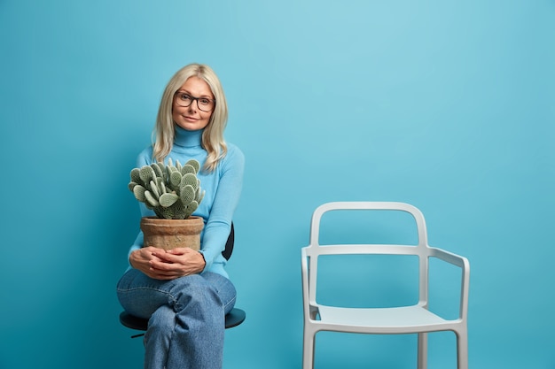 beautiful blonde woman with European appearance holds pot of cactus sits alone near empty chair being on self isolation at home needs live communication