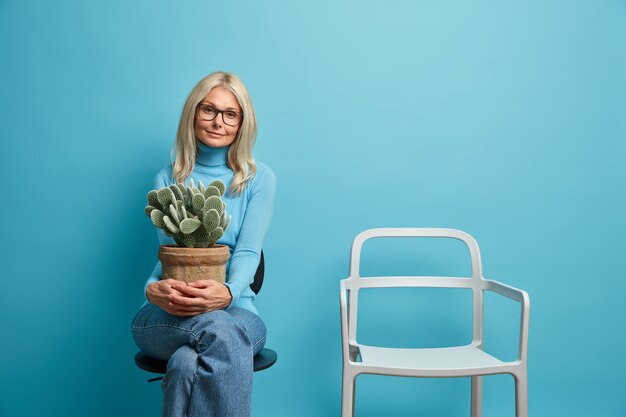 beautiful blonde woman with European appearance holds pot of cactus sits alone near empty chair being on self isolation at home needs live communication