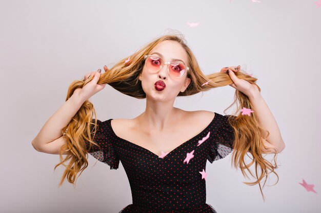 Beautiful blonde woman with long curly hair in hands having fun, cheerful girl giving kiss, looking happy. Wearing pink glasses, black pretty dress. Pink confetti stars. Isolated..