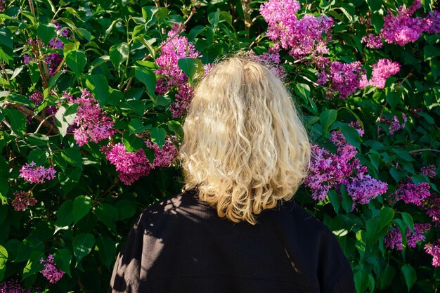 黒いデニムのジャケットを着た長いウェーブのかかった髪の美しいブロンドの女性は、ライラックの茂みの咲く庭に立っています春の時間女性は田舎のライラック公園で休む春の自然を楽しんでいます