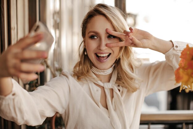 Beautiful blonde young woman in pearl necklace and stylish white blouse shows peace sign smiles and takes selfie in restaurant