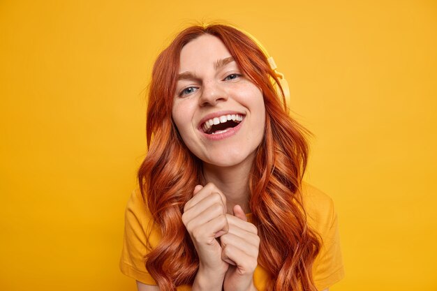 Beautiful blue eyed cheerful woman has red wavy hair keeps hands together smiles broadly enjoys leisure time listens music in stereo headphones with good sound quality 