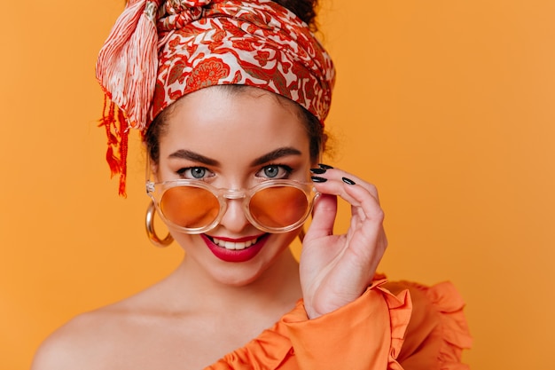 Foto gratuita bella signora dagli occhi azzurri in insolita fascia e rossetto rosso si toglie gli occhiali da sole e posa sullo spazio arancione.