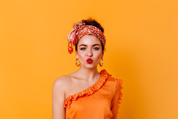 Beautiful blue-eyed woman in orange silk outfit and stylish headband blows air kiss on isolated space.