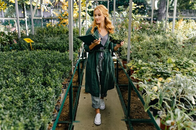 Beautiful botanical garden, filled with many green flowers and bushes. Girl with blond curly hair, poses, presenting herself as biologist.