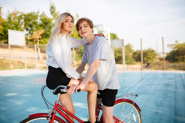 Beautiful boy and girl with blond hair on bicycle happily looking in camera while spending time together on basketball court Young cute couple riding on red bicycle in park