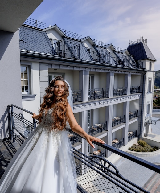 Foto gratuita la bella sposa si è vestita in vestito bianco di lusso sul balcone di un appartamento il giorno soleggiato